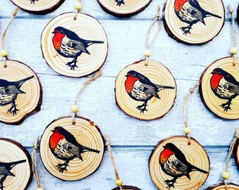 May include: A set of 12 wooden ornaments featuring a hand-painted robin design. Each ornament is a round slice of wood with a rustic finish and a red, black, and white robin illustration. The ornaments are strung with twine and have a small wooden bead at the end.