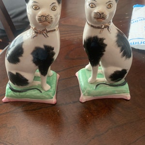 May include: Pair of black and white porcelain cat figurines sitting on green and pink cushions. The cats have gold accents around their eyes and collars.