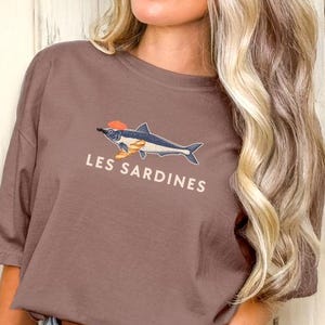 May include: A brown t-shirt with a graphic of a fish with a red hat and the text "Les Sardines" printed on the front.