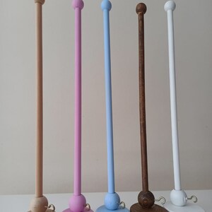 May include: Five wooden dowel rods with rounded tops and bases. The dowels are painted in natural wood, pink, light blue, dark brown, and white. Each dowel has a small gold hook attached to the base.