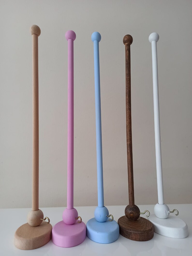 May include: Five wooden dowel rods with rounded tops and bases. The dowels are painted in natural wood, pink, light blue, dark brown, and white. Each dowel has a small gold hook attached to the base.