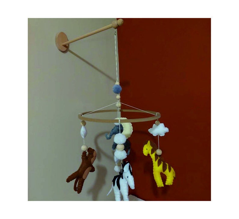 May include: A wooden mobile with felt animal toys hanging from a circular wooden frame. The animals include a zebra, giraffe, elephant, and a brown animal. The mobile is suspended from a wooden arm attached to the wall.
