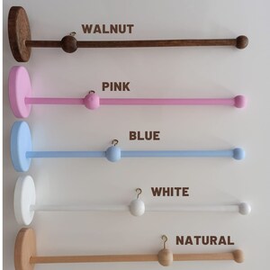 May include: Five wooden baby mobile hangers in different colors: walnut, pink, blue, white, and natural. Each hanger has a round base and a long, straight bar with a hook at one end and a wooden ball at the other.