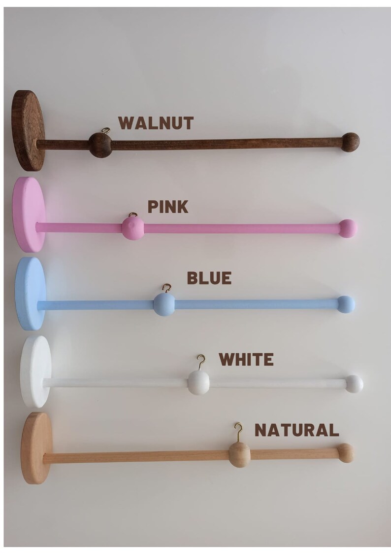 May include: Five wooden baby mobile hangers in different colors: walnut, pink, blue, white, and natural. Each hanger has a round base and a long, straight bar with a hook at one end and a wooden ball at the other.