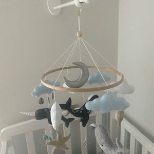 May include: A white wooden mobile arm attached to a white wall with a wooden ring mobile hanging from it. The mobile features a crescent moon, clouds, a narwhal, a whale, and a starfish. The mobile is hanging over a white crib.