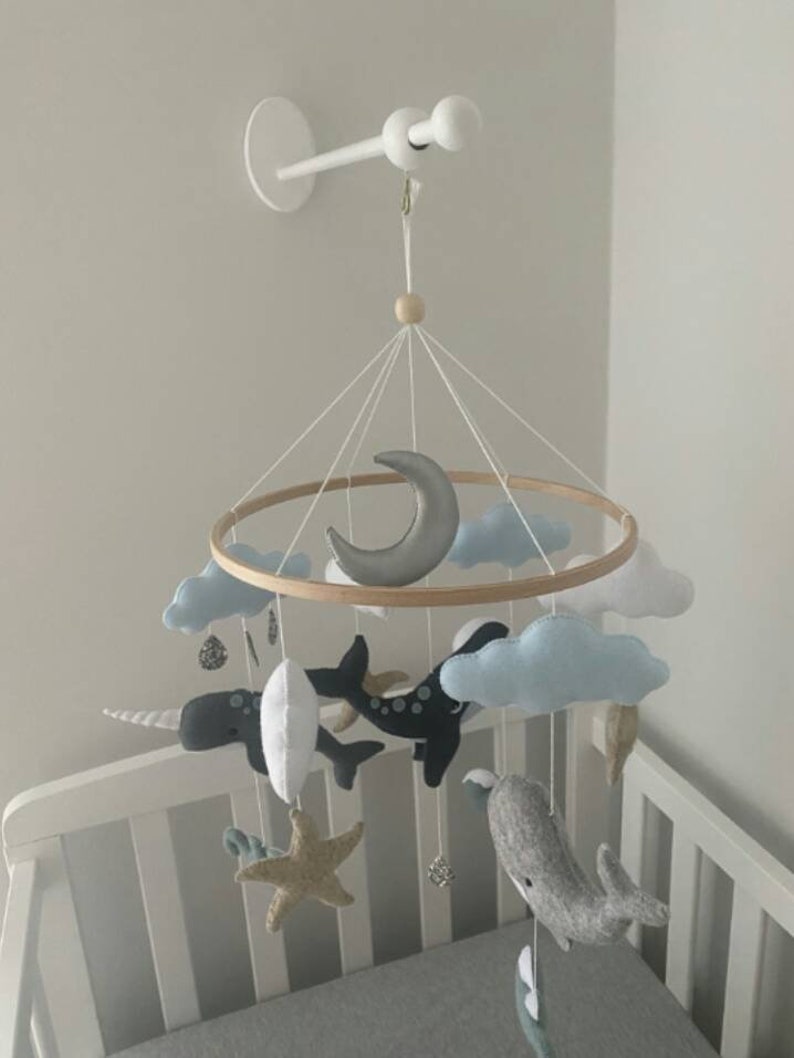 May include: A white wooden mobile arm attached to a white wall with a wooden ring mobile hanging from it. The mobile features a crescent moon, clouds, a narwhal, a whale, and a starfish. The mobile is hanging over a white crib.