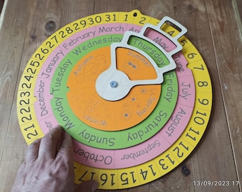 May include: A wooden calendar with a rotating dial to show the days of the week, months, and seasons. The calendar is yellow, pink, green, and orange. The dial is labeled with the days of the week, months, and seasons. The numbers 1 through 31 are on the outer ring.
