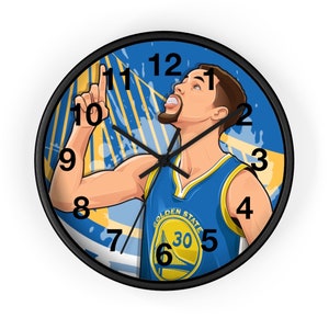 May include: Round wall clock with a black frame. The clock face features a cartoon illustration of a basketball player in a blue and gold jersey, pointing upwards. The clock hands are black, and the numbers are in a black, bold font.