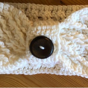 May include: Cream-coloured, hand-knitted headband with a cable knit design. The headband features a large, dark brown button for closure. The soft, textured yarn creates a cosy and stylish accessory.