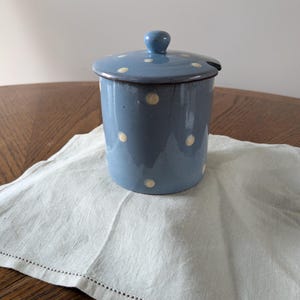 Può includere: Un barattolo di zucchero in ceramica blu con un coperchio bianco e pois bianchi. Il barattolo è appoggiato su un tovagliolo di lino bianco.