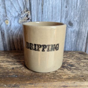 Puede incluir: Un recipiente de cerámica beige con la palabra "DRIPPING" en letras negras. El recipiente es cilíndrico con un borde ligeramente ensanchado. Se encuentra sobre una estantería de madera contra un fondo de madera envejecida.