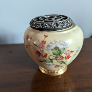 Peut inclure: Un vase en céramique de couleur crème avec un couvercle en métal argenté et un motif floral. Le vase a une base dorée et est décoré de fleurs roses et rouges, de feuilles vertes et d'accents blancs.