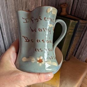 Pode incluir: Um jarro de cerâmica azul-claro com uma pega curva e um bico. O exterior apresenta um texto castanho pintado à mão que diz "I fye can't be as I be as I can't be as I been 181" e um pequeno motivo floral com pétalas brancas e centro castanho.