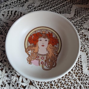 May include: White ceramic bowl with an Art Nouveau style design. It features a woman with auburn hair and a floral headdress, framed by a gold and brown border. The bowl is a decorative item.