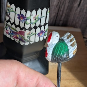May include: A decorative pin depicting a small, colourful bird with a white head, red beak, and green body. The bird has gold and white accents. The pin is held in front of a black vase with a floral and bird design.