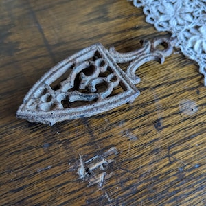 Puede incluir: Un salvamanteles de hierro fundido oxidado con un diseño calado ornamentado. El salvamanteles es triangular con un asa y se encuentra sobre una superficie de madera. El diseño presenta patrones arremolinados.