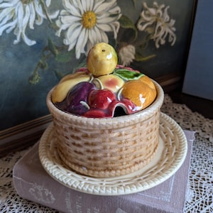Op de afbeelding: Een keramische mandvormige container met een deksel versierd met kleurrijk fruit. De container staat op een bijpassend bord, geplaatst bovenop een boek. De achtergrond toont een schilderij van witte bloemen.