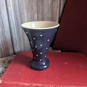 Könnte beinhalten: Eine kleine, marineblaue Keramikvase mit weißen Tupfen. Die Vase hat eine ausgestellte Oberseite und einen abgerundeten Boden. Der Innenraum ist cremefarben. Die Vase steht auf einem roten Buch.