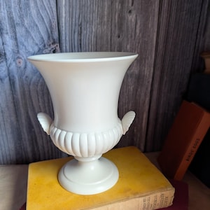 Op de afbeelding: Een matte, gebroken witte keramische vaas in urnstijl met een brede uitlopende rand, een geribbelde onderkant en twee kleine schelpvormige handvatten aan de zijkanten. De vaas rust op een ronde voet en heeft een gladde, egale afwerking.