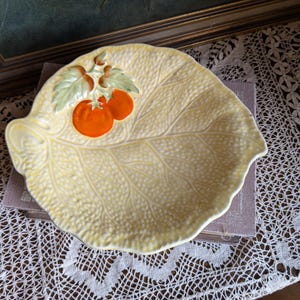 Könnte beinhalten: Eine blattförmige, gelbe Keramikschale mit strukturierter Oberfläche. Die Schale zeigt eine Ansammlung von drei orangen Tomaten mit grünen Blättern. Die Schale ist wahrscheinlich vintage und eignet sich zum Servieren oder Ausstellen.