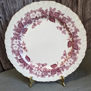 May include: A white ceramic dinner plate with a scalloped edge and a purple floral design. The design features a wreath of blackberry vines with leaves and flowers.