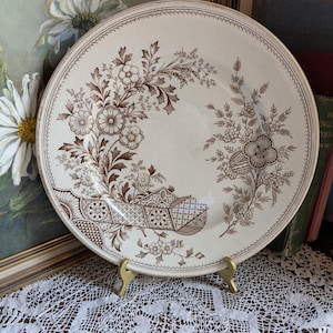 Peut inclure: Une assiette antique crème avec un motif floral marron. L'assiette présente un motif détaillé de fleurs, de feuilles et de formes géométriques. Le bord a un bord festonné. L'assiette est exposée sur un support doré.