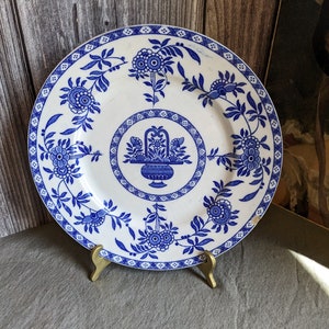 Può includere: Un piatto in ceramica bianca con un motivo floreale blu e bianco. Il piatto ha un bordo rialzato e un disegno centrale di un cesto di fiori. Il piatto è su un supporto in metallo dorato.