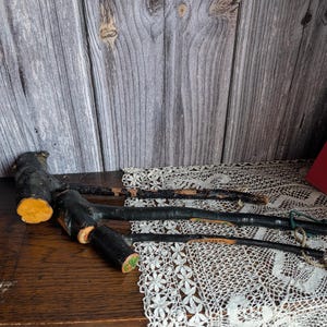 Könnte beinhalten: Mehrere rustikale Wanderstöcke mit schwarz lackierten Abschnitten und natürlichen Holzakzenten. Die Stöcke sind auf einer Holzoberfläche mit einer weißen Spitzendecke angeordnet. Die Enden der Stöcke haben eine gelb-orange Farbe.