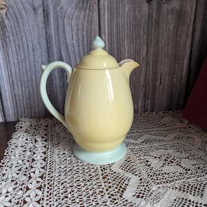 Può includere: Una teiera vintage in ceramica gialla con base, manico e pomolo del coperchio azzurri. La teiera ha un bordo dorato e un beccuccio curvo. È appoggiata su una tovaglia di pizzo bianco.