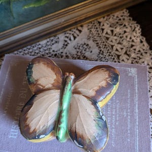Può includere: Una statuetta di farfalla in ceramica dipinta di marrone, giallo e verde. La farfalla ha un motivo dettagliato sulle ali e un corpo verde.