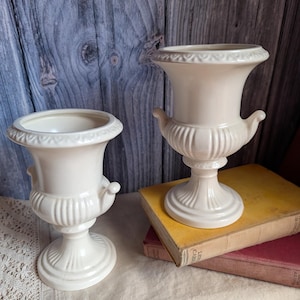 Op de afbeelding: Twee crèmekleurige keramische vazen in urnstijl met voetstuk, uitlopende randen met een decoratief reliëfpatroon en kleine handvatten aan de zijkant. De vazen hebben een verticaal geribbeld ontwerp op het onderste deel van de behuizing.