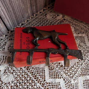 May include: Bronze-coloured metal wall hook featuring a dog silhouette above a rifle design, with four hooks below. The hook is resting on a red book titled "The Observer's Book of Dogs". The background includes a wooden surface and a white lace tablecloth.