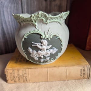 Peut inclure: Un pot en céramique vert pâle orné d'un médaillon en forme de cœur vert foncé avec un relief blanc représentant un chérubin jouant de la flûte parmi des nuages. Le bord présente une finition festonnée ornementale dans un ton vert plus clair.