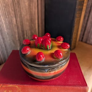 May include: A ceramic trinket box with a lid, decorated with red cherries on top. The box has a black, orange, and brown color scheme. The box is sitting on top of a red book.