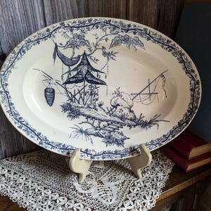 May include: A large oval white ceramic serving platter with a blue and white floral design. The design features a pagoda, a tree, and a basket hanging from a branch. The platter is on a white stand.