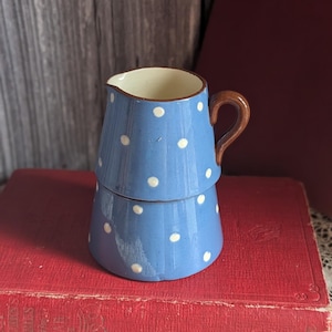 Puede incluir: Una pequeña jarra de cerámica azul con lunares blancos. Tiene un asa y borde marrón. La jarra está colocada sobre un libro rojo. Una adición encantadora a cualquier cocina o comedor.