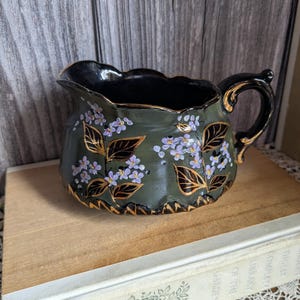 Peut inclure: Petite cruche en céramique vert foncé avec intérieur noir et bordure dorée. La cruche est ornée de fleurs violettes et d'accents de feuilles d'or. Elle a une anse et repose sur une surface en bois.
