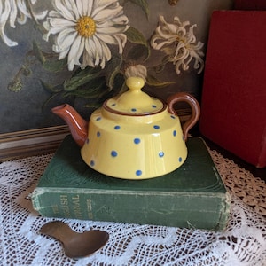 Peut inclure: Une théière en céramique jaune à pois bleus, avec une anse marron et un bec verseur rouge. La théière est posée sur un livre vert, avec une cuillère en bois et une peinture florale en arrière-plan. Un bel objet pour le thé.
