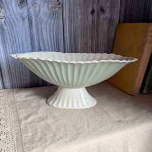 Puede incluir: Un cuenco de pedestal de cerámica verde pálido con un borde festoneado y un diseño exterior acanalado. El cuenco se apoya sobre una base acampanada a juego, presentando una amplia abertura de forma ovalada con un acabado liso y brillante.