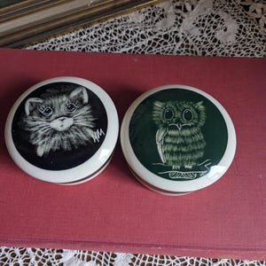Può includere: Due scatole rotonde in ceramica per piccoli oggetti con coperchi. Una scatola presenta un'illustrazione in bianco e nero di un gatto con occhi verdi. L'altra scatola presenta uno sfondo verde con un'illustrazione bianca di un gufo con occhi gialli.