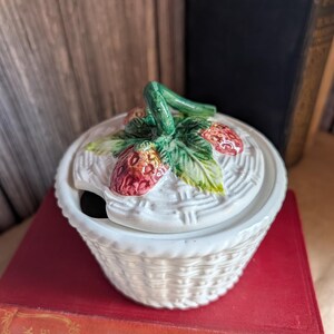 Peut inclure: Pot à confiture en céramique blanche en forme de panier, avec un couvercle orné de fraises. Le couvercle présente une tige et des feuilles vertes avec des fraises rouges. Le pot est posé sur un livre rouge.