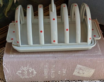 May include: A vintage white metal toast rack with red polka dots. The rack has six arched slots and a tray base. The design is simple and functional, perfect for serving toast. The rack is sitting on a stack of books.