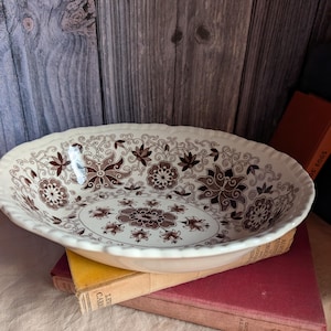 May include: An oval ceramic bowl with a scalloped edge, featuring a detailed brown floral and botanical pattern printed on a cream-coloured background. The interior of the bowl displays a repeating design of stylised flowers and leaves.