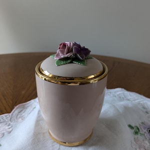 Puede incluir: Un tarro de cerámica rosa con un borde dorado y una tapa floral. La tapa presenta una rosa rosa y morada con hojas verdes.