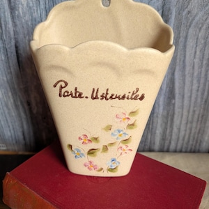 May include: A beige ceramic wall pocket with a scalloped top and a hole for hanging. The front is decorated with hand-painted flowers in pink and blue, with the words "Porte. Ustensiles" in brown script.