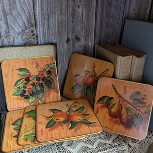 Può includere: Una collezione di sottobicchieri quadrati in legno con illustrazioni di frutta. I sottobicchieri presentano immagini di ciliegie, pesche e pere su uno sfondo di venature del legno. I sottobicchieri sono disposti su una tovaglia di pizzo bianco.