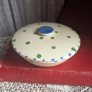 May include: A cream-coloured ceramic bowl with a lid, decorated with green and blue polka dots. The lid has a small blue knob. The bowl is sitting on a red book.