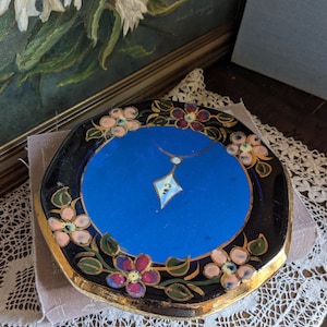 Op de afbeelding: Achtekantige sieradendoos met een blauwe binnenkant en gouden rand. Het deksel heeft een geschilderd ontwerp van roze en rode bloemen met gouden accenten en een zilveren ketting met een diamanten hanger.