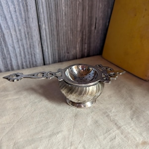 May include: A silver-toned metal tea strainer featuring a bowl with a perforated interior and a decorative handle with cut-out details. The base of the strainer has a rounded, swirling ribbed design, and the handle extends horizontally from the side.