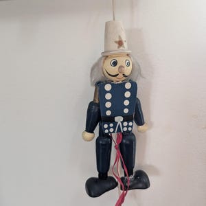 Peut inclure: Ornement de soldat en bois avec un chapeau haut de forme blanc orné d'une étoile rouge. Le soldat porte un uniforme bleu avec des boutons blancs et une moustache grise. Des rubans rouges et blancs pendent du bas. Un objet décoratif.